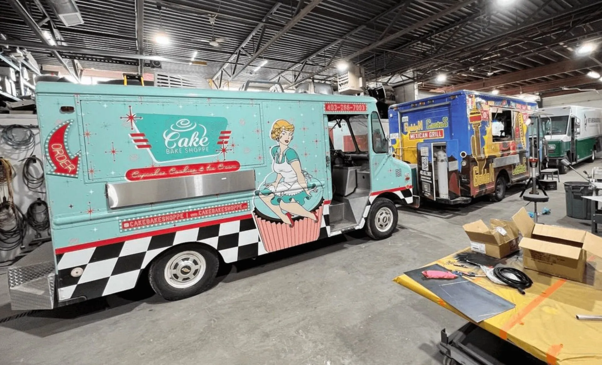 Retro-themed cake food truck with a checkerboard stripe, in the shop with other trucks